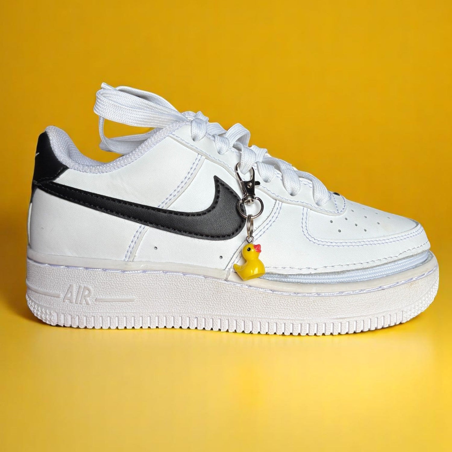 White Nike Air Force sneaker with black swoosh logo and yellow rubber duck charm on a yellow background