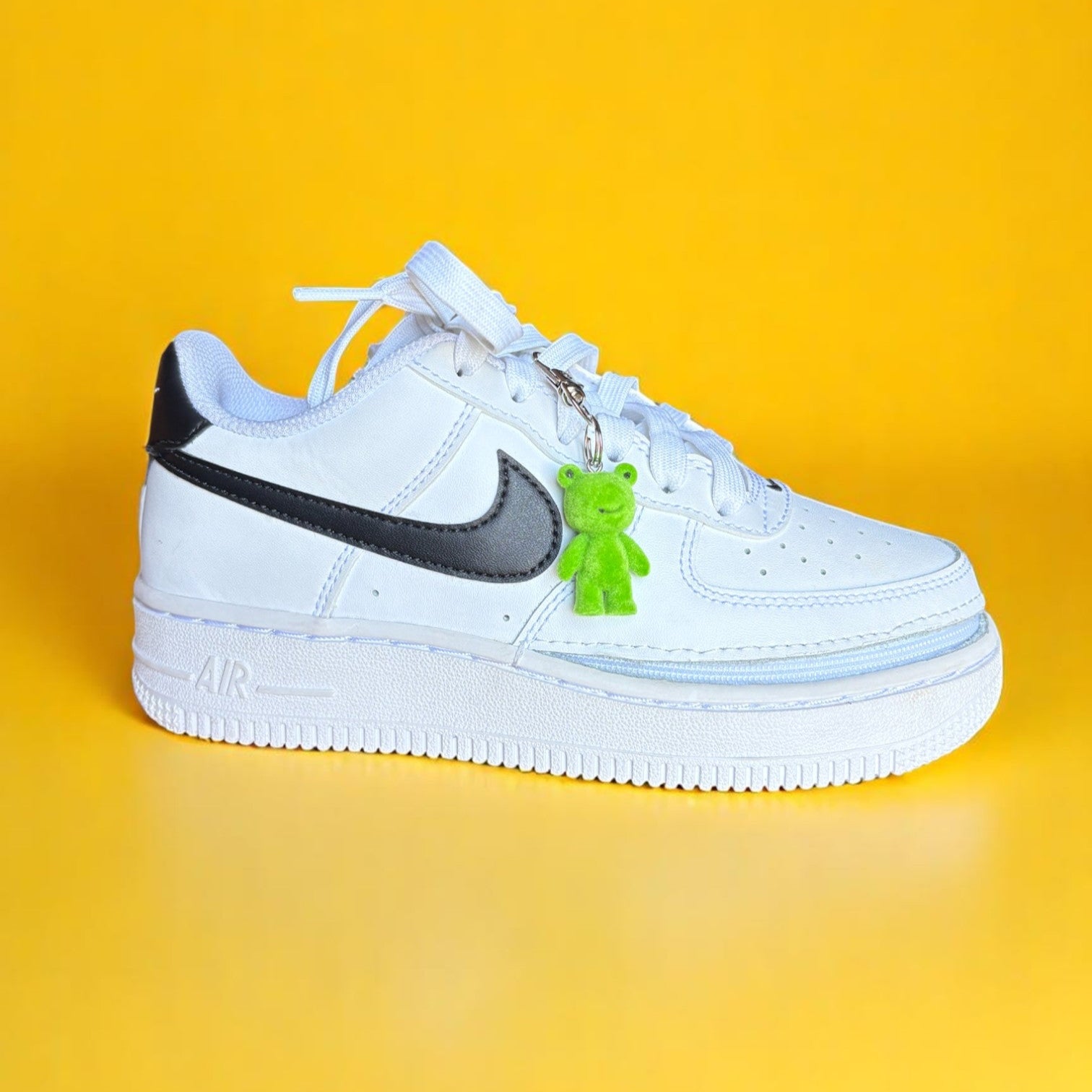 White sneaker with black swoosh and green frog charm on a yellow background